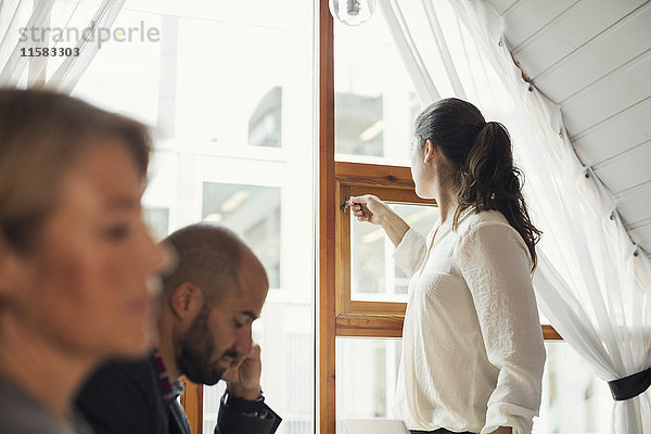 Mittlere erwachsene Geschäftsfrau schließt Bürofenster  während Kollegen im Vordergrund stehen