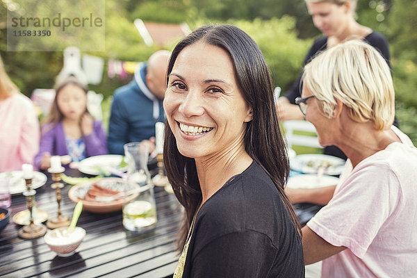 Porträt einer glücklichen Frau  die mit Familie und Freunden am Esstisch im Garten sitzt.