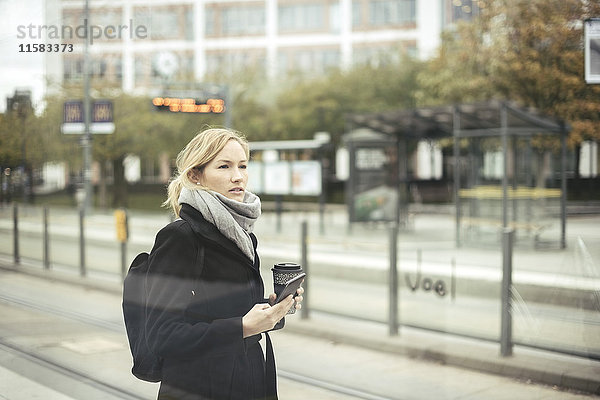 Mittlere erwachsene Geschäftsfrau mit Handy und Einweg-Kaffeetasse an der Straßenbahnhaltestelle