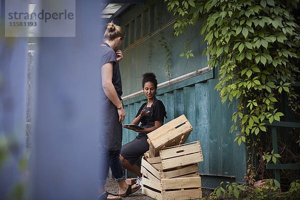 Gärtnerin hält digitales Tablett und kommuniziert mit einem Freund über Kisten im Garten.