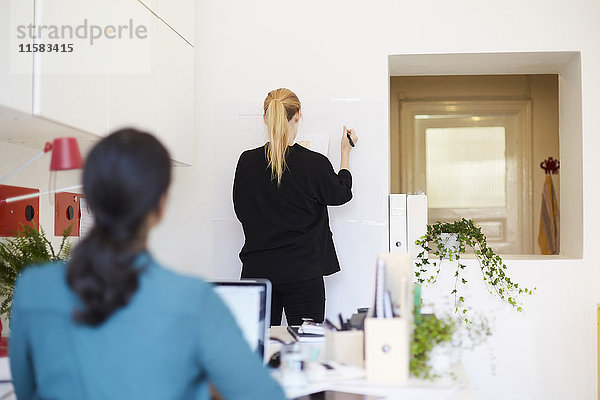 Rückansicht der Geschäftsfrau beim Schreiben auf Whiteboard mit Kollegin im Vordergrund im Büro