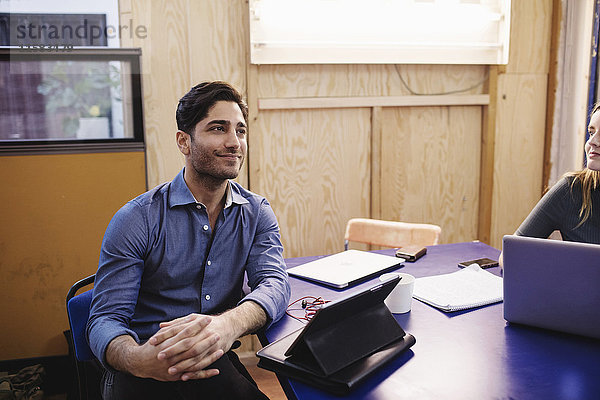Lächelnder junger Mann schaut weg  während er im Büro am Schreibtisch sitzt.