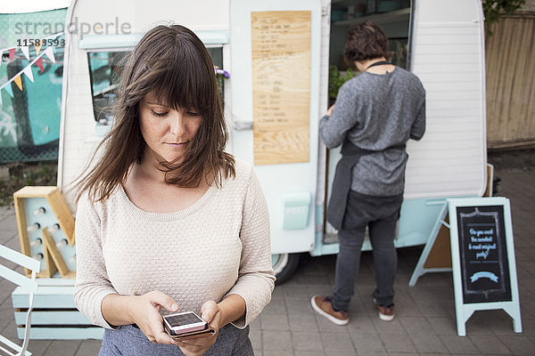 Besitzerin nutzt Smartphone außerhalb des Lebensmittelwagens  während die Kollegin im Hintergrund arbeitet.