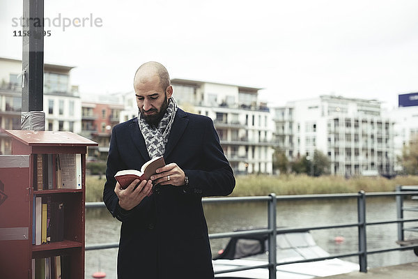 Mittlerer erwachsener Geschäftsmann beim Lesen eines Buches am Fluss in der Stadt