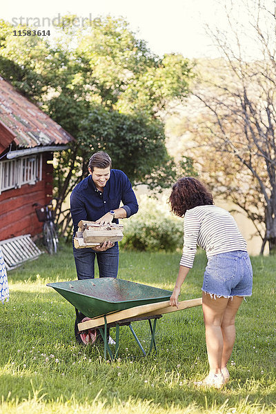 Mann stellt Brennholz  während die Frau an einem sonnigen Tag eine Schubkarre auf dem Hof hält.