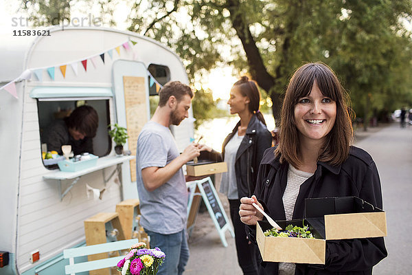 Lächelnder Kunde mit Einweg-Salatschachtel auf der Straße mit Freunden und Besitzer außerhalb des Food-Trucks im Hintergrund