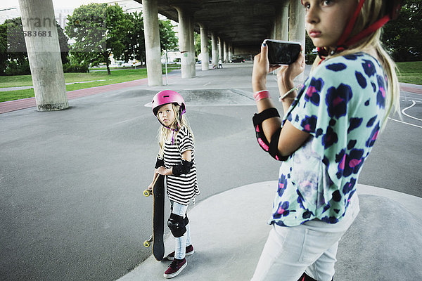 Mädchen fotografiert Freundin mit Skateboard durch Handy im Park