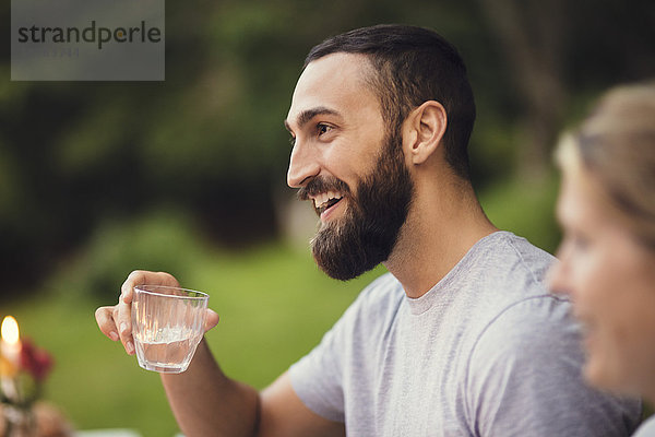 Lächelnder Mann hält Trinkglas  während er mit einem Freund auf einer Gartenparty sitzt.