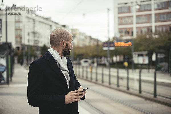 Mittlerer Erwachsener Geschäftsmann hält Smartphone  während er auf dem Bürgersteig in der Stadt steht.