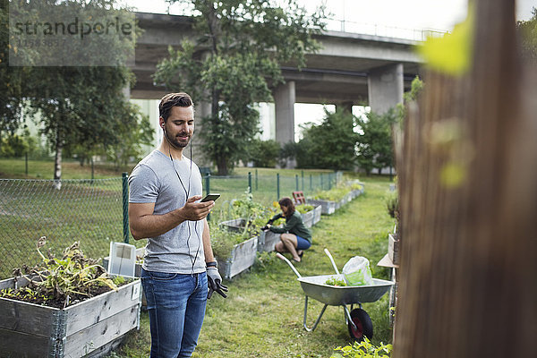 Mittlerer erwachsener Mann mit Handy  während Frauen im Hintergrund im Stadtgarten arbeiten.