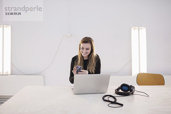 Glückliche Bloggerin mit Smartphone am Schreibtisch im Büro