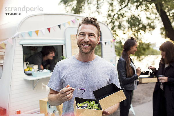 Lächelnder männlicher Kunde  der mit Freunden und Besitzer im Hintergrund eine Einweg-Salatschachtel gegen den Essenswagen hält.