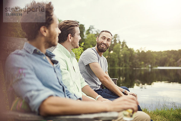Fröhliche männliche Freunde beim Sitzen auf einer Holzbank am Seeufer