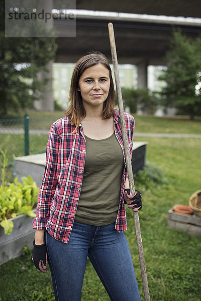 Porträt einer mittleren erwachsenen Frau mit Rechen im Gemeinschaftsgarten