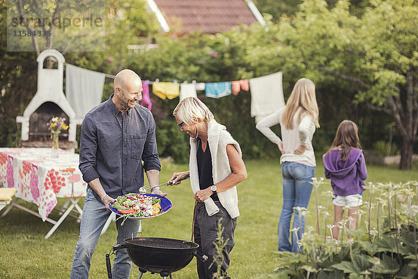 Mann und Frau kochen Gemüse auf dem Grill mit Mädchen  die während der Gartenparty im Garten stehen.