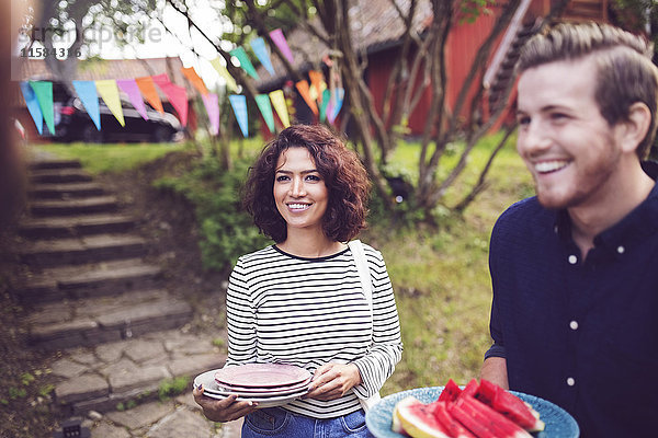 Lächelnde Freunde halten Teller  während sie während der Gartenparty im Garten stehen.