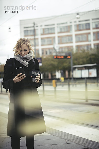 Mittlere erwachsene Geschäftsfrau  die ein Handy benutzt  während sie eine Einweg-Kaffeetasse auf dem Bürgersteig in der Stadt hält.