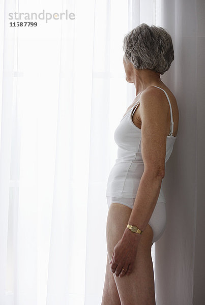 Seniorin in weißer Unterwäsche am Fenster stehend