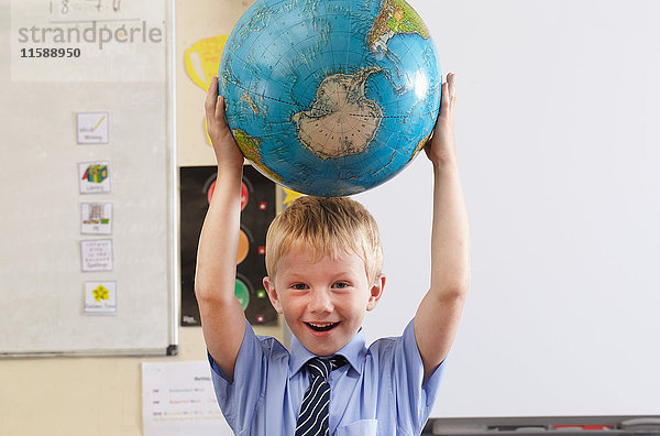 Schuljunge  der in einem Klassenzimmer einen Globus über seinen Kopf hält