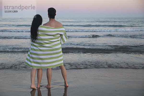 Rückansicht des jungen Paares am Strand in Handtuch gehüllt  um den Sonnenuntergang zu beobachten.