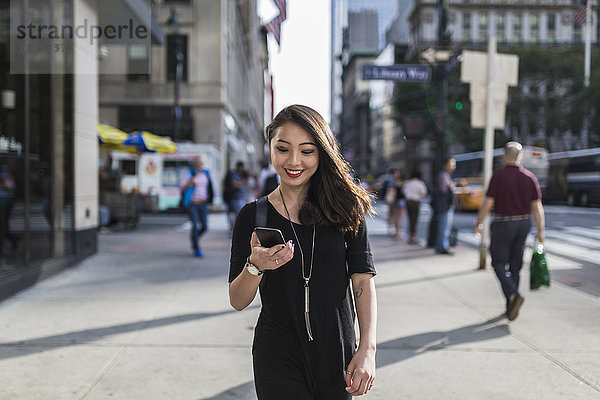 USA  New York City  Manhattan  Porträt einer lächelnden jungen Frau in Schwarz mit Blick auf das Handy