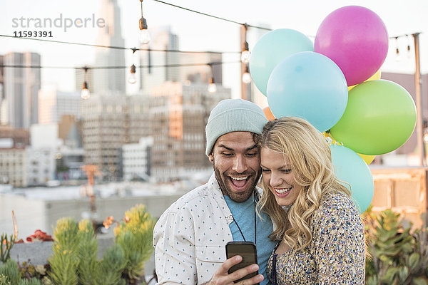 Junges Paar mit Luftballons auf der Dachterrasse mit Smartphone