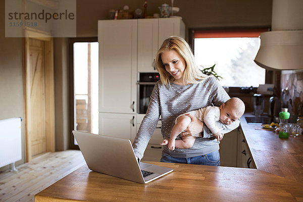 Lächelnde Mutter mit Baby in der Küche mit Laptop