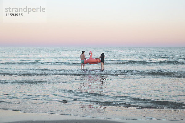 Rückansicht des jungen Paares  das mit aufblasbarem rosa Flamingo ins Meer geht