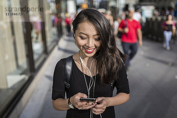 USA  New York City  Manhattan  junge Frau  die mit Kopfhörern Musik hört  während sie auf ihr Handy schaut.
