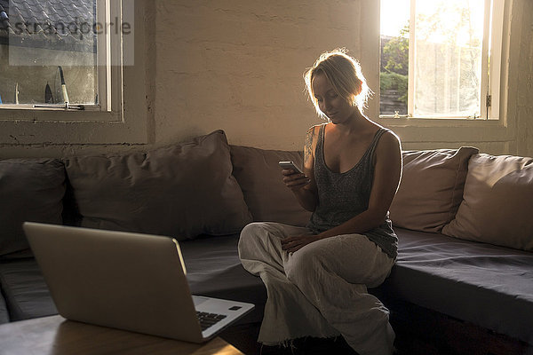 Blonde Frau sitzt auf der Couch bei Gegenlicht und schaut aufs Handy.