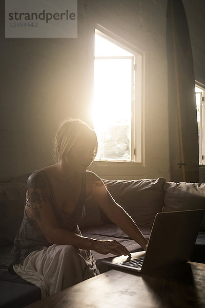 Blonde Frau sitzt auf der Couch bei Gegenlicht mit Laptop
