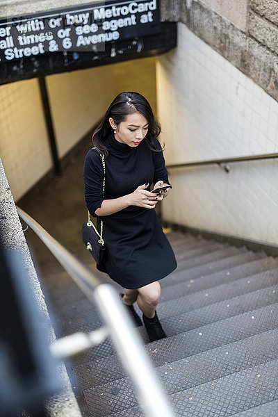 USA  New York City  Manhattan  junge Frau in schwarz gekleidet  die nach oben geht und das Handy ansieht.