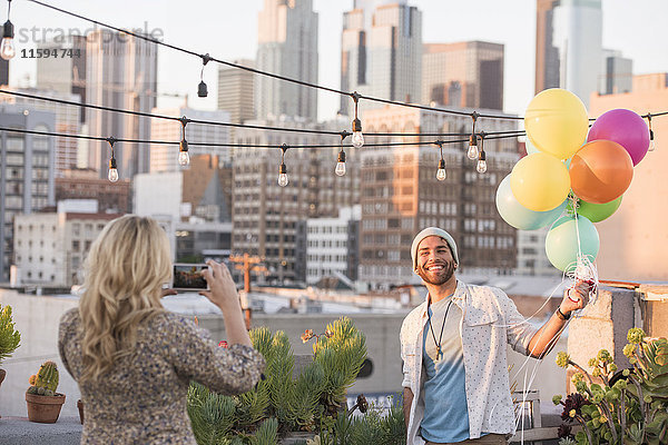 Junges Paar mit Luftballons auf der Dachterrasse mit Smartphone