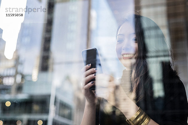 USA  New York City  Manhattan  lächelnde junge Frau hinter Glasscheibe beim Blick aufs Handy