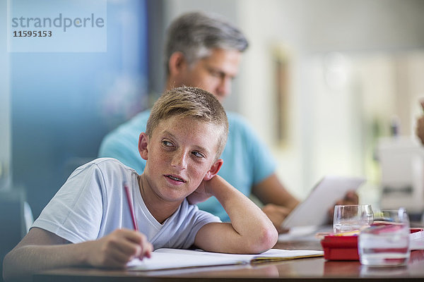 Junge macht Hausaufgaben mit Vater im Hintergrund