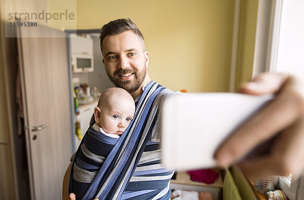 Vater mit Babysohn im Tragetuch zu Hause mit einem Selfie