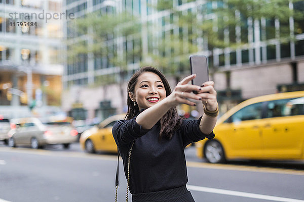 USA  New York City  Manhattan  Porträt einer lächelnden jungen Frau  die sich selbst mit dem Smartphone fotografiert.