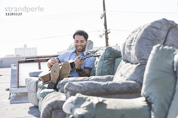 Junger Mann auf dem Dach sitzt auf dem Sofa und spielt Gitarre.