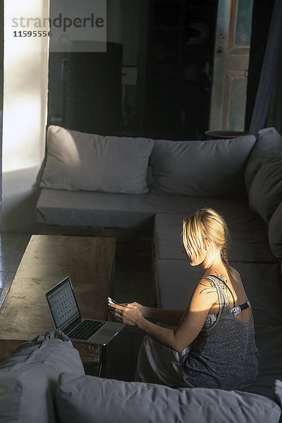 Blonde Frau sitzt auf der Couch bei Sonnenlicht mit dem Handy