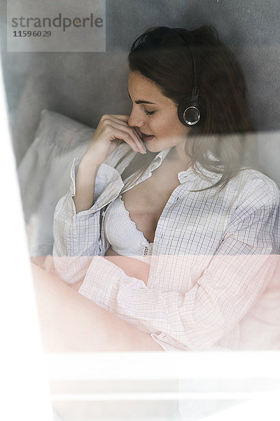 Junge Frau hinter der Fensterscheibe Musik hören mit Kopfhörern zu Hause