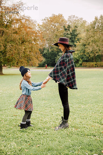 Junge Frau und kleines Mädchen stehen sich im Herbst auf einer Wiese gegenüber und halten sich an den Händen.