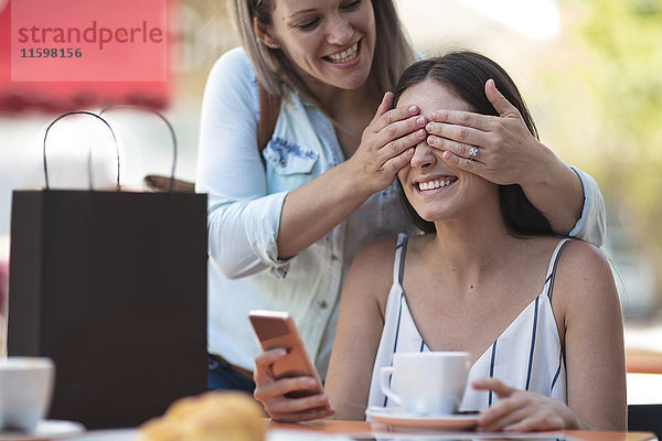 Frau bedeckt die Augen ihrer Freundin in einem Outdoor-Café
