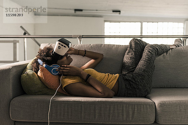 Junge Frau auf Couch liegend mit Kopfhörer und VR-Brille