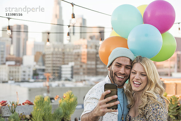 Junges Paar mit Luftballons auf der Dachterrasse mit Smartphone