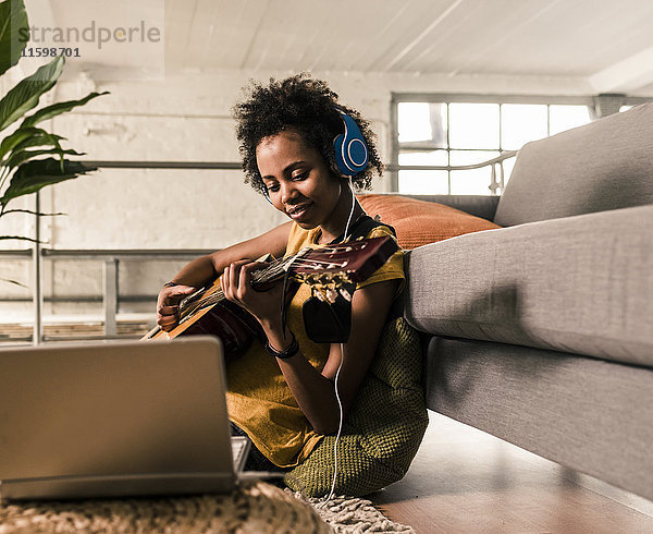 Junge Frau zu Hause mit Kopfhörer und Laptop beim Gitarrespielen