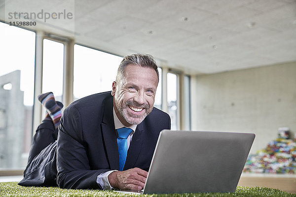 Glücklicher Geschäftsmann liegt auf Kunstrasen mit Laptop
