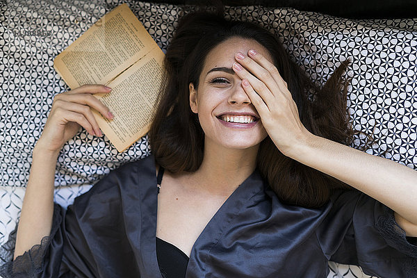 Porträt einer lächelnden jungen Frau auf dem Bett liegend mit einem Buch  das ein Auge mit der Hand bedeckt.