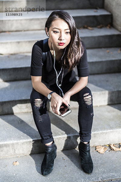 Porträt einer jungen Frau in Schwarz  die auf einer Treppe sitzt und Musik mit Kopfhörer und Handy hört.