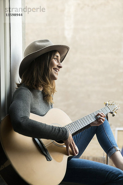 Lächelnde junge Frau sitzt im Fensterrahmen und spielt Gitarre.