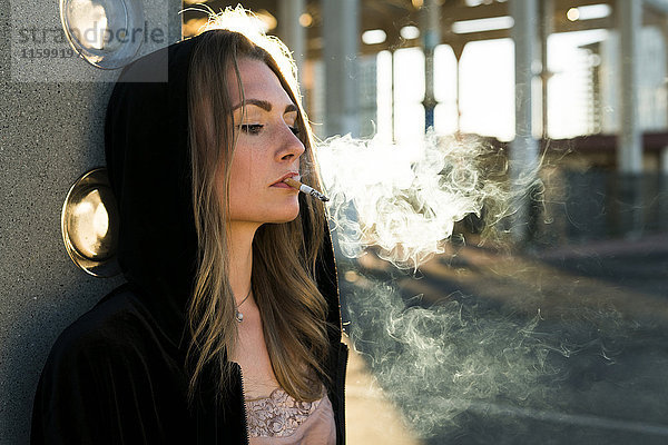 Porträt einer jungen Frau in Kapuzenjacke mit Zigarette im Gegenlicht